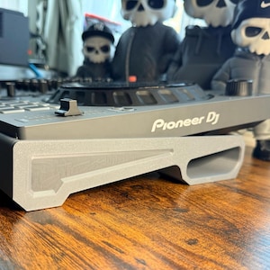 Pode incluir: Um controlador Pioneer DJ cinza com vários botões e botões. O controlador está em uma superfície de madeira. Ao fundo, há várias figuras em forma de caveira usando chapéus e jaquetas.