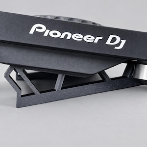 Pode incluir: Um controlador Pioneer DJ preto montado num suporte angular preto. O dispositivo apresenta um acabamento preto mate com o logótipo branco Pioneer DJ impresso na lateral. Dois pés circulares prateados são visíveis na parte inferior.