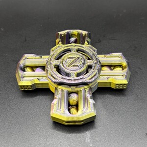 May include: A multi-colored fidget spinner with a cross-like design. The spinner features a central circular element with the letter "Z" in the center, surrounded by a decorative pattern. The arms of the spinner are yellow and purple.