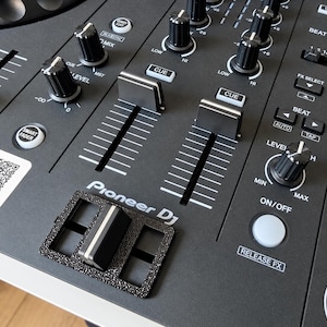 Peut inclure: Un contrôleur DJ Pioneer noir doté d'un cache de crossfader noir texturé, de deux faders de canal verticaux, de plusieurs boutons rotatifs et de divers boutons de commande avec texte blanc, dont Beat FX, Cue et Smart Fader, avec un autocollant QR.