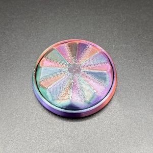 Puede incluir: Un objeto redondo y multicolor con una superficie texturizada. El objeto presenta un diseño circular con líneas radiales en tonos morado, rosa, azul y naranja. El objeto está sobre un fondo gris oscuro.