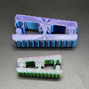 Könnte beinhalten: Zwei 3D-gedruckte Objekte sind abgebildet. Das größere ist blau und lila, das kleinere grün und rosa. Beide haben eine strukturierte Oberfläche und ein einzigartiges, geometrisches Design. Es handelt sich wahrscheinlich um dekorative oder funktionale Gegenstände.