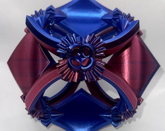 Könnte beinhalten: Eine geometrische Skulptur in Blau und Burgunderrot. Das komplexe Design zeigt rautenförmige und gebogene Elemente, die symmetrisch angeordnet sind. Aufwendige Details sind sichtbar und schaffen ein visuell ansprechendes Kunstwerk.
