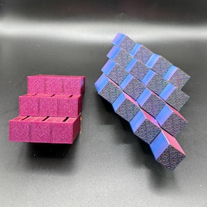 Può includere: Due oggetti geometrici stampati in 3D. Uno è una pila di tre blocchi rettangolari color magenta. L'altro è una serie di cubi blu e magenta iridescenti collegati, che formano una forma flessibile a forma di serpente. Gli oggetti sono su una superficie scura.