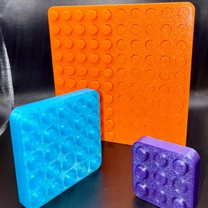 May include: A set of building blocks in orange, blue, and purple. The set includes a large orange square, a blue square, and a smaller purple square. Each piece has a textured surface with circular studs.
