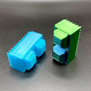 Könnte beinhalten: Zwei 3D-gedruckte geometrische Formen. Eine ist eine leuchtend blaue, eckige Form mit gezackter Oberseite und zwei abgerundeten Vorsprüngen. Die andere ist eine grün-blaue Stapelstruktur mit strukturiertem Muster und gezackter Oberseite.