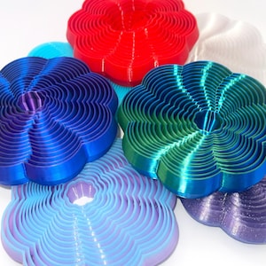 Puede incluir: Conjunto de seis cortadores de galletas con forma de flor en varios colores, incluyendo rojo, azul, verde, morado y blanco. Cada cortador tiene múltiples capas, creando un efecto de capas.