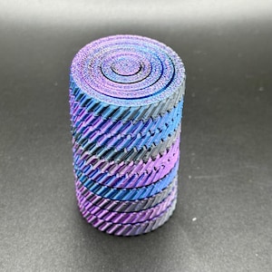 Könnte beinhalten: Ein zylindrisches Objekt mit einer spiralförmigen Oberseite und einem strukturierten, geschichteten Design. Die Farben wechseln von Blau zu Lila und Grau und erzeugen einen schillernden Effekt. Das Objekt steht aufrecht auf einer dunklen Oberfläche.