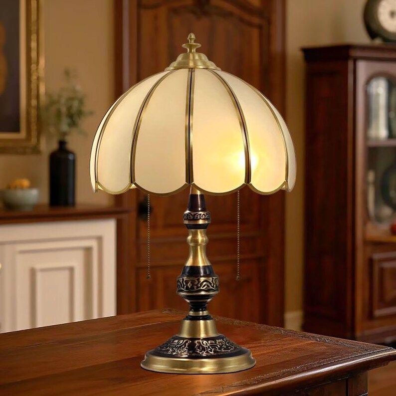 Pu&ograve; includere: Una lampada da tavolo in stile vintage con paralume a conchiglia color crema e dettagli in ottone. La base presenta dettagli ornati e una finitura marrone scuro e dorata. La lampada ha due catenelle ed &egrave; su un tavolo di legno.