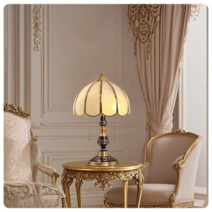 Pu&ograve; includere: Una lampada da tavolo in stile vintage con paralume a conchiglia color crema e base in bronzo scuro. La lampada &egrave; posizionata su un tavolino dorato ornato. Il design della lampada presenta un'estetica classica ed elegante.