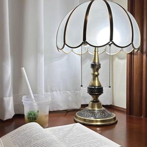 Pu&ograve; includere: Una lampada da tavolo in stile vintage con paralume bianco smerlato e base color ottone. La lampada &egrave; su una superficie di legno scuro accanto a un libro aperto e una bevanda in un bicchiere trasparente.