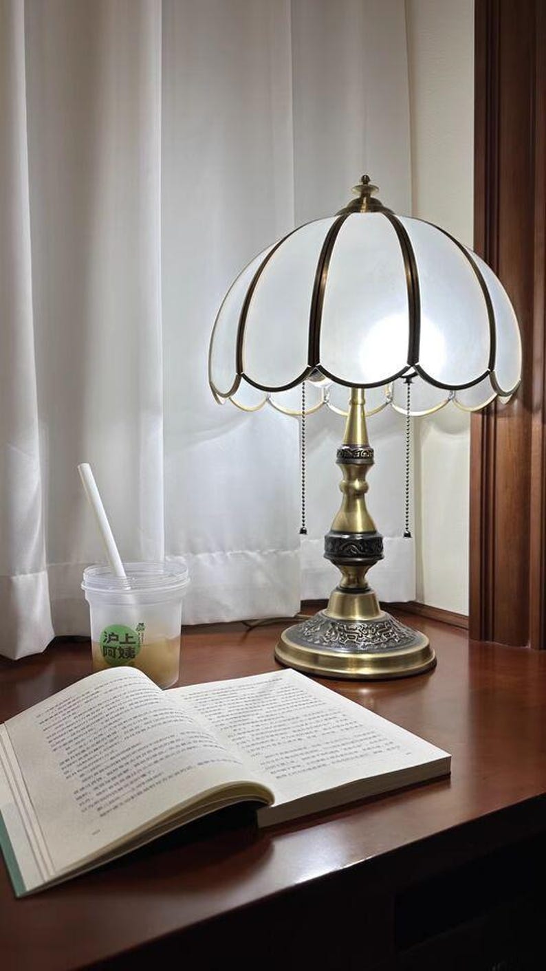 Pu&ograve; includere: Una lampada da tavolo in stile vintage con paralume bianco smerlato e base color ottone. La lampada &egrave; su una superficie di legno scuro accanto a un libro aperto e una bevanda in un bicchiere trasparente.