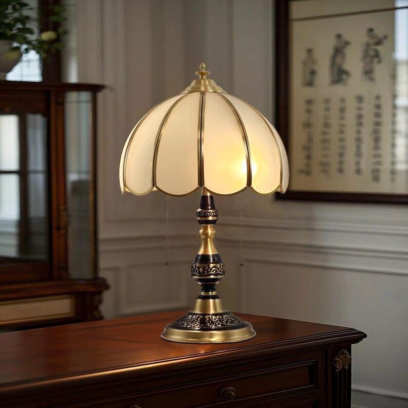 Pu&ograve; includere: Una lampada da tavolo in stile vintage con paralume a conchiglia color crema e base in ottone. La lampada presenta un design decorativo e ornato sulla base e sullo stelo. La lampada &egrave; accesa, emettendo un bagliore caldo e invitante.