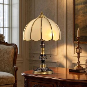 Pu&ograve; includere: Una lampada da tavolo in stile vintage con paralume a conchiglia color crema e dettagli in ottone. La lampada presenta una base decorativa e uno stelo dettagliato. &Egrave; su un tavolo di legno, con un candeliere sullo sfondo.