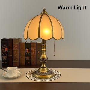 Pu&ograve; includere: Una lampada da tavolo in stile vintage con luce calda, caratterizzata da un paralume smerlato color crema e una base in ottone. La lampada &egrave; posizionata davanti a una fila di libri antichi. Il testo "Warm Light" &egrave; visibile sopra la lampada.