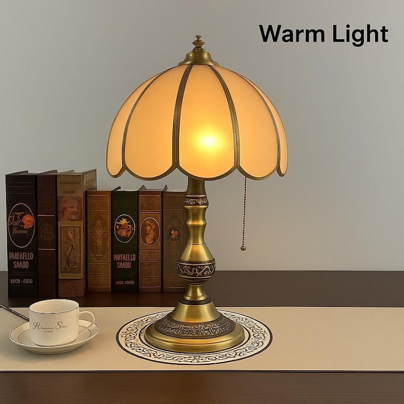 Pu&ograve; includere: Una lampada da tavolo in stile vintage con luce calda, caratterizzata da un paralume smerlato color crema e una base in ottone. La lampada &egrave; posizionata davanti a una fila di libri antichi. Il testo "Warm Light" &egrave; visibile sopra la lampada.