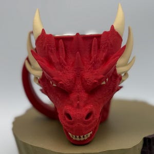 May include: A red dragon-shaped mug with cream-colored horns and teeth. The mug has a detailed, textured surface, and a red handle. The mug is designed to resemble a dragon's head, with a menacing expression.