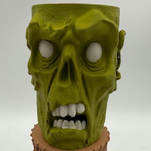 May include: A green zombie head-shaped container with white eyes and a wide open mouth revealing white teeth. The container is made of a textured material and sits on a brown base. The design is detailed and grotesque.