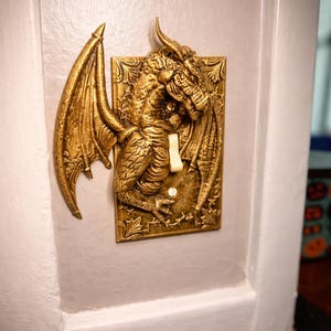 May include: A gold-colored dragon-themed light switch cover. The switch plate features a detailed relief of a dragon with outstretched wings. The switch itself is a standard toggle style, and the plate is mounted on a white wall.