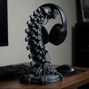May include: A dark gray, spine-shaped headphone stand. The stand has a mechanical design with gears and simulated vertebrae. A pair of black headphones rests on the stand. The stand is on a wooden desk with a keyboard and mouse.