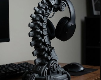 May include: A dark gray, spine-shaped headphone stand. The stand has a mechanical design with gears and simulated vertebrae. A pair of black headphones rests on the stand. The stand is on a wooden desk with a keyboard and mouse.