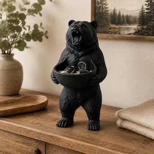 May include: A matte black bear figurine standing on its hind legs with its mouth open in a roar. The bear holds a matching black bowl in its front paws, which contains a set of metal keys and several small coins, serving as a functional tray.