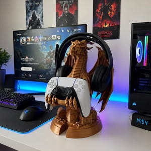 May include: A brown dragon-shaped gaming headset and controller stand. The stand holds a black headset and a white and black controller. The stand is on a white desk with a computer monitor and keyboard in the background.