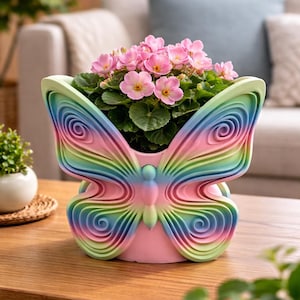 May include: A butterfly-shaped planter with a rainbow-colored wing design. The planter is filled with pink flowering plants. The planter is on a wooden table.