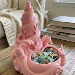 May include: A pink decorative sculpture featuring a swirling tower, seahorse figures, and wave-like details. The sculpture holds a collection of sea glass, seashells, and starfish, creating a coastal theme.
