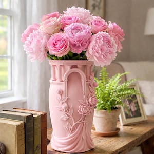 May include: A pink decorative vase filled with pink peonies and roses. The vase has an architectural design with columns and floral relief. The arrangement sits on a wooden table with old books and a potted fern.