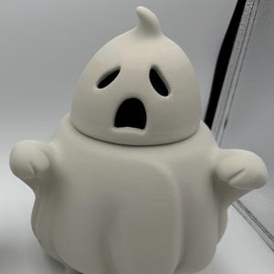 May include: A white ceramic ghost-shaped container with a lid. The ghost has a surprised expression with open eyes and mouth. The container has small arms and feet, perfect for Halloween decor.