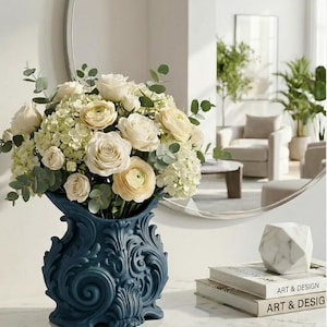May include: A navy blue, ornate vase filled with white roses, hydrangeas, and eucalyptus. The vase has a detailed, swirling design. The arrangement sits on a white surface, with a marble sculpture and books labeled "ART & DESIGN" nearby.