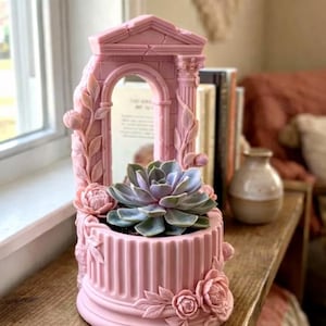 May include: A pink planter with an architectural design, featuring a central archway and fluted base. The planter is adorned with floral and leaf details and holds a succulent plant with green and purple leaves. A decorative piece for home decor.