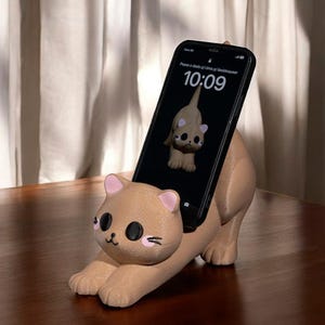 May include: A tan cat-shaped phone holder with pink accents, holding a black smartphone. The phone displays a cat image and the time 10:09. The holder is on a wooden surface.