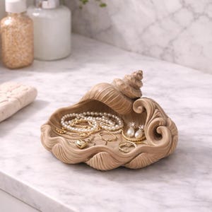 May include: A beige seashell-shaped jewelry tray filled with necklaces, rings, and earrings. The tray is made of a textured material and sits on a marble countertop. The jewelry includes gold and pearl accents.