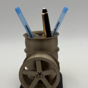 May include: A 3D-printed pen holder in the shape of a car wheel and barrel. The holder is tan and holds three pens: two blue and one black and gold. The base is black.