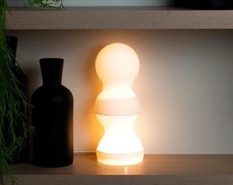 May include: A modern, glowing table lamp with a white, rounded shade and a wooden band. The lamp is on a wooden shelf, next to a black vase and greenery. The lamp emits a warm, inviting light.