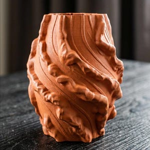 May include: A terracotta-colored vase with a textured, organic design. The vase has a swirling, layered appearance with a rough, uneven surface. The open top suggests it is designed to hold flowers or other decorative items. The vase is sitting on a dark wooden surface.