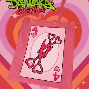 May include: Pink tote bag with a pink and white playing card design. The card is the four of hearts and features a pair of scissors with heart-shaped handles. The text "DAYWAKA CUSTOMS" is visible in the top left corner of the image.