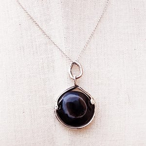Può includere: Una collana d'argento con un grande pendente rotondo in pietra nera. Il pendente è incastonato in una lunetta d'argento e pende da una delicata catena d'argento. La pietra preziosa ha una superficie lucida e riflettente.
