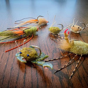 May include: Assortment of six hand-tied fishing flies, featuring a variety of colors and materials. The flies are designed to resemble crabs and other crustaceans.