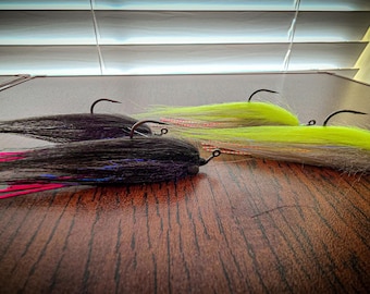 May include: Two fishing lures with metal hooks. One lure features long black hair with pink accents, while the other has bright neon yellow hair, a grey body, and a white eye. Both lures are designed for fly fishing and have a streamlined shape.
