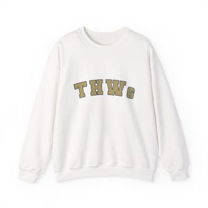 Puede incluir: Una sudadera blanca de cuello redondo con las letras "THWG" en una fuente de bloque dorada y marrón en el pecho. La sudadera tiene mangas largas y un diseño clásico e informal.