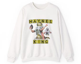 Pode incluir: Camisola branca com um gráfico de um jogador de futebol americano em uniforme dourado e o texto "HAYNES KING" em letras amarelas. O design inclui uma segunda imagem do jogador. A camisola é de gola redonda.