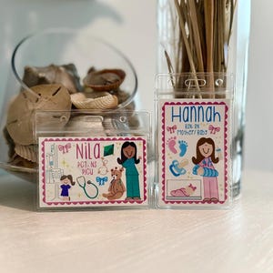 May include: Two clear plastic name badge holders with colorful illustrations. One says "Nila" with medical-themed graphics, and the other says "Hannah" with baby-related images. Both have pink borders and are on a white surface.