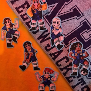 Op de afbeelding: Een verzameling stickers met cartoon illustraties van voetbalsters in blauw-gouden uniformen. De stickers zijn geplaatst op een oranje achtergrond en een grijze stof met de tekst "YELLOW JACKETS".