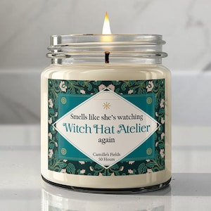 May include: A lit candle in a clear glass jar with a label featuring a teal and dark green floral pattern. The label text reads: Smells like she's watching Witch Hat Atelier again, Camille's Fields, 50 Hours. The wax is off-white and the flame is bright.