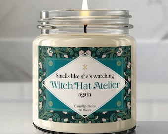 May include: A lit candle in a clear glass jar with a label featuring a teal and dark green floral pattern. The label text reads: Smells like she's watching Witch Hat Atelier again, Camille's Fields, 50 Hours. The wax is off-white.