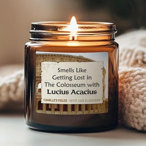 May include: A lit candle in a brown glass jar. The label reads "Smells Like Getting Lost in The Colosseum with Lucius Acacius." The candle is from Camille's Fields, with white sage and lavender scents.