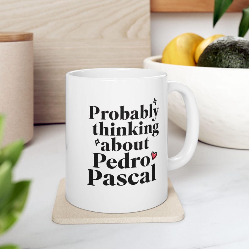 Puede incluir: Taza de cer&aacute;mica blanca con el texto "Probably thinking about Pedro Pascal" en negro. La taza tiene un asa blanca y est&aacute; sobre un posavasos beige. Un s&iacute;mbolo de coraz&oacute;n est&aacute; al lado del nombre.